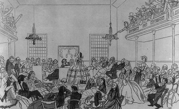 A black and white 19th-century engraving illustrates a woman delivering a speech in a large, crowded hall, likely at an early women's rights convention. The speaker, wearing a voluminous dress, stands centrally before a seated audience that includes many women, while a balcony filled with men shows several reacting with apparent hostility and jeers.